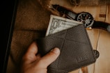 A hand holding a dark leather wallet with a one-dollar bill partially visible. A rustic background includes a burlap sack, a wristwatch with a black leather strap, and a couple of coins placed on a wooden surface.