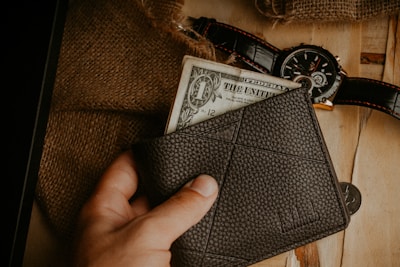 A hand holding a dark leather wallet with a one-dollar bill partially visible. A rustic background includes a burlap sack, a wristwatch with a black leather strap, and a couple of coins placed on a wooden surface.