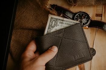A hand holding a dark leather wallet with a one-dollar bill partially visible. A rustic background includes a burlap sack, a wristwatch with a black leather strap, and a couple of coins placed on a wooden surface.