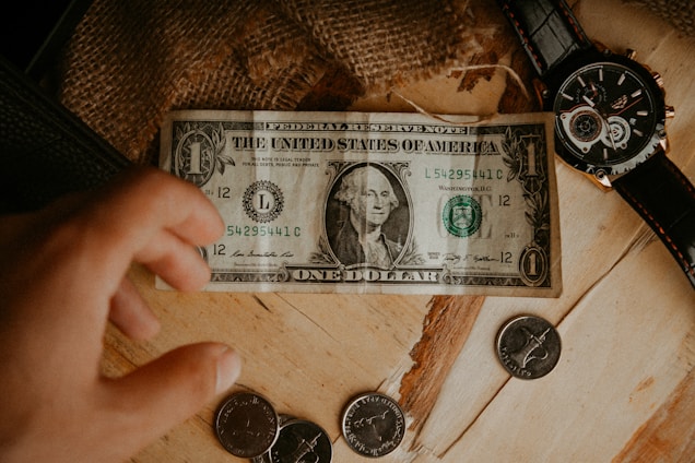 A hand reaches towards coins and a one dollar bill placed on a wooden surface. A black wristwatch with a round face is positioned to the side. The background includes a rustic textured fabric.