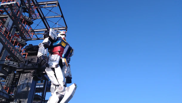 A large robot structure stands adjacent to a metal framework. The robot is designed with colorful panels, including red, blue, yellow, and white sections, giving it a futuristic and mechanical appearance. The background is a clear blue sky, creating a stark contrast with the detailed features of the robot and structure.