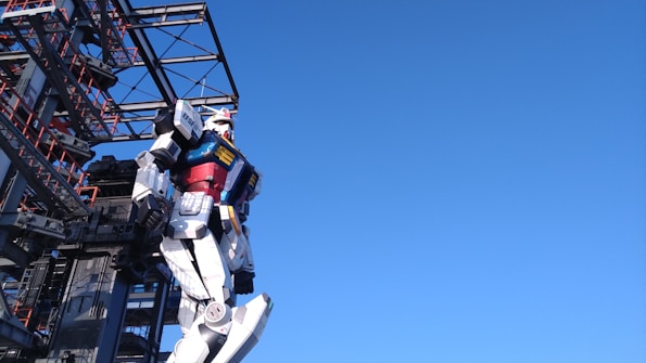 A large robot structure stands adjacent to a metal framework. The robot is designed with colorful panels, including red, blue, yellow, and white sections, giving it a futuristic and mechanical appearance. The background is a clear blue sky, creating a stark contrast with the detailed features of the robot and structure.