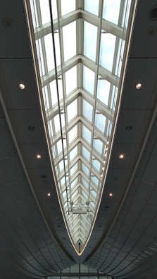 A modern architectural ceiling design with large triangular glass panels, framed by metal structures, creating an elongated and symmetrical visual effect. Ambient lighting is integrated along the edges, highlighting the sleek and futuristic aesthetic.