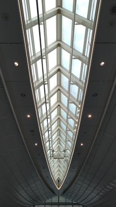 A modern architectural ceiling design with large triangular glass panels, framed by metal structures, creating an elongated and symmetrical visual effect. Ambient lighting is integrated along the edges, highlighting the sleek and futuristic aesthetic.