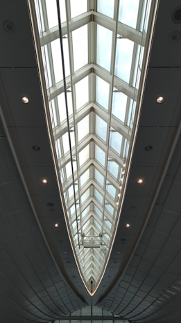 A modern architectural ceiling design with large triangular glass panels, framed by metal structures, creating an elongated and symmetrical visual effect. Ambient lighting is integrated along the edges, highlighting the sleek and futuristic aesthetic.