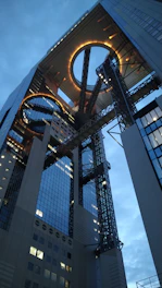 An architectural structure featuring two high-rise buildings connected at the top with circular openings and illuminated edges. The design incorporates modern glass and steel elements with an industrial aesthetic.