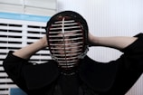 A person is wearing a kendo helmet and protective gear, adjusting the helmet with both hands behind their head. The setting appears to be indoors, with white and blue slatted blinds in the background.