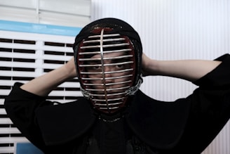 A person is wearing a kendo helmet and protective gear, adjusting the helmet with both hands behind their head. The setting appears to be indoors, with white and blue slatted blinds in the background.