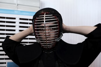A person is wearing a kendo helmet and protective gear, adjusting the helmet with both hands behind their head. The setting appears to be indoors, with white and blue slatted blinds in the background.