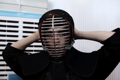 A person is wearing a kendo helmet and protective gear, adjusting the helmet with both hands behind their head. The setting appears to be indoors, with white and blue slatted blinds in the background.