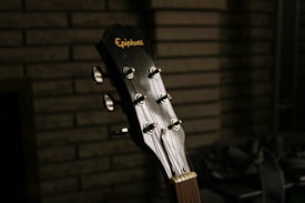 A close-up view of a guitar headstock with tuning pegs visible. The brand name is engraved on the headstock, and the background consists of brick or block patterns, adding a textured backdrop.