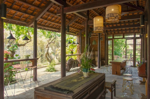 A cozy, rustic outdoor patio merges with an interior space through large glass doors. The setting features wooden beams, stone flooring, and lush greenery. Several tables and stools are placed throughout, complemented by hanging light fixtures that create a warm ambiance. Plants and decorative items add charm to the scene.