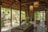 A cozy, rustic outdoor patio merges with an interior space through large glass doors. The setting features wooden beams, stone flooring, and lush greenery. Several tables and stools are placed throughout, complemented by hanging light fixtures that create a warm ambiance. Plants and decorative items add charm to the scene.