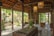 A cozy, rustic outdoor patio merges with an interior space through large glass doors. The setting features wooden beams, stone flooring, and lush greenery. Several tables and stools are placed throughout, complemented by hanging light fixtures that create a warm ambiance. Plants and decorative items add charm to the scene.