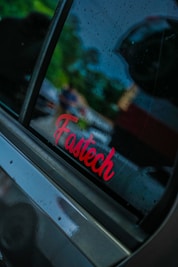A car window with raindrops and a red 'Fastech' sticker. The background shows blurred greenery and some indistinct shapes, suggesting a rainy or overcast day.