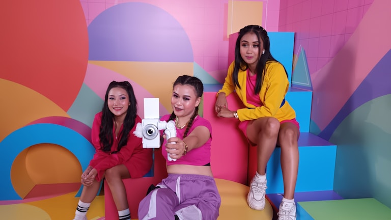 Three women are posing in a brightly colored, geometric-themed room. They are dressed in vibrant clothing, with one holding a camera-like object and the others sitting on colorful steps. The background features bold shapes and a palette of pastels and primary colors.