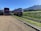 A double-decker bus is parked on a gravel lot next to a colorful tram on railway tracks. The setting is in a rural area with a grassy hill and mountain range in the background under a clear blue sky.