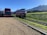 A double-decker bus is parked on a gravel lot next to a colorful tram on railway tracks. The setting is in a rural area with a grassy hill and mountain range in the background under a clear blue sky.