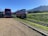 A double-decker bus is parked on a gravel lot next to a colorful tram on railway tracks. The setting is in a rural area with a grassy hill and mountain range in the background under a clear blue sky.