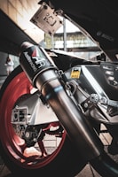 A close-up view of a motorcycle exhaust system with branding visible, highlighting the sleek metallic design against a background of red wheels and logos.
