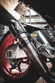 A close-up view of a motorcycle exhaust system with branding visible, highlighting the sleek metallic design against a background of red wheels and logos.
