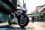 A sleek, sporty motorcycle with a glossy finish is parked on an urban street. The motorcycle displays a bold design with red, white, and blue accents. The sun is setting, casting a warm glow over the cityscape, and tall buildings surround the area. A person walks in the background, contributing to the bustling city atmosphere.