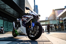 A sleek, sporty motorcycle with a glossy finish is parked on an urban street. The motorcycle displays a bold design with red, white, and blue accents. The sun is setting, casting a warm glow over the cityscape, and tall buildings surround the area. A person walks in the background, contributing to the bustling city atmosphere.