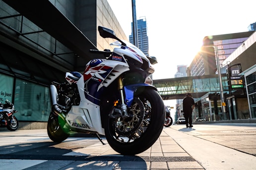 A sleek, sporty motorcycle with a glossy finish is parked on an urban street. The motorcycle displays a bold design with red, white, and blue accents. The sun is setting, casting a warm glow over the cityscape, and tall buildings surround the area. A person walks in the background, contributing to the bustling city atmosphere.