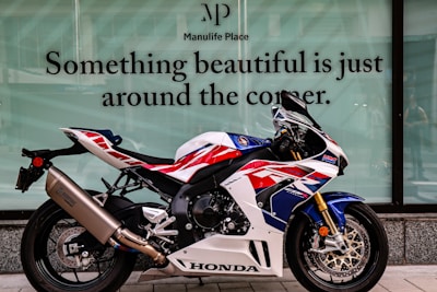 A sporty Honda motorcycle is parked on a sidewalk in front of a large glass window. The motorcycle features a distinct color scheme with red, blue, and white accents. On the window behind it, there is a motivational quote written in large letters. The background is composed of a modern building facade.