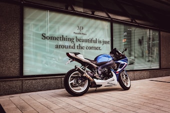 A blue and white Suzuki motorcycle is parked on a tiled pathway in front of a large glass window displaying the phrase 'Something beautiful is just around the corner.' The setting appears to be an urban environment with modern architecture.