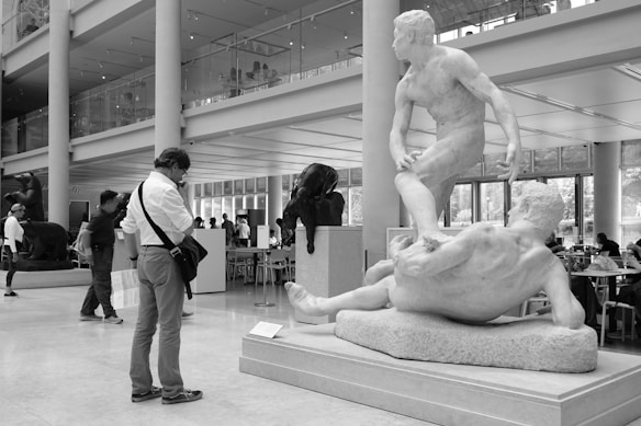 A museum or gallery space with a large classical sculpture depicting two figures in dynamic poses. People are observing the art, and the setting includes modern architectural elements like pillars and glass walls. The atmosphere combines artistic appreciation with leisure, as there are tables and people sitting in the background.