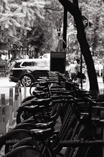 A row of bicycles is parked at a bike rental station, with a stop sign visible in the background. Trees and foliage surround the area, creating a natural setting. Several people are walking on the sidewalk, and a black car is parked near the bike station. The image is in black and white, giving it a classic and calm feel.