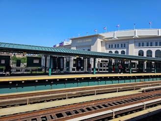 A train station platform with a large building in the background displaying flags on its roof. Several people are standing on the platform, near the 161st Street sign. The tracks are visible in the foreground.