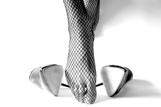Fishnet stockings are worn on legs poised atop large, reflective, triangular high heels. The image has a minimalist aesthetic with a focus on texture and contrast.