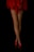 Fishnet stockings cover crossed legs, complemented by red high heels and a flowing red fabric skirt against a black background.