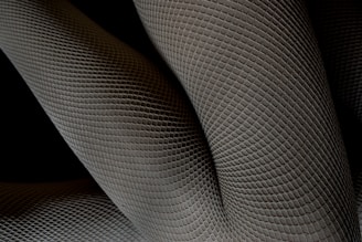 A close-up of textured fabric with a geometric pattern, appearing as overlapping layers with a three-dimensional effect. The pattern resembles scales or a mesh, creating an intricate and abstract design.