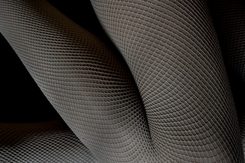 A close-up of textured fabric with a geometric pattern, appearing as overlapping layers with a three-dimensional effect. The pattern resembles scales or a mesh, creating an intricate and abstract design.