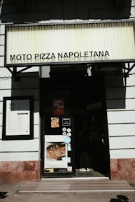 The image shows the entrance of a pizza restaurant named Moto Pizza Napoletana. There is a large sign with the restaurant's name at the top. The door has various stickers and a sign showing the opening hours. On the left is a menu board mounted on the wall. A poster displaying a pizza is also visible on the door.