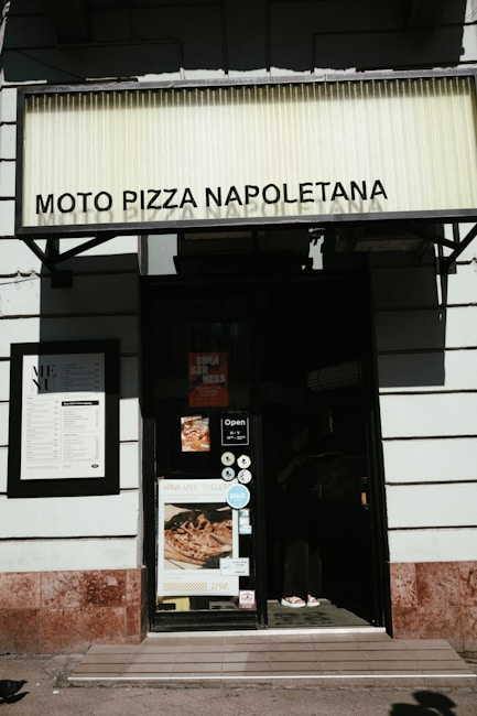 The image shows the entrance of a pizza restaurant named Moto Pizza Napoletana. There is a large sign with the restaurant's name at the top. The door has various stickers and a sign showing the opening hours. On the left is a menu board mounted on the wall. A poster displaying a pizza is also visible on the door.