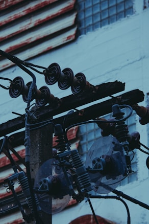 A close-up view of electrical components and wires mounted on a utility pole, with insulators and transformers visible. The background shows a building with a mix of red and white painted surfaces and glass block windows.