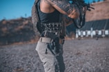 An individual with tattoos on their arm is wearing a tactical vest and holding a firearm. They are standing in an outdoor setting, possibly a shooting range, with targets visible in the background.