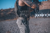 An individual with tattoos on their arm is wearing a tactical vest and holding a firearm. They are standing in an outdoor setting, possibly a shooting range, with targets visible in the background.