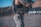 An individual with tattoos on their arm is wearing a tactical vest and holding a firearm. They are standing in an outdoor setting, possibly a shooting range, with targets visible in the background.