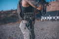 An individual with tattoos on their arm is wearing a tactical vest and holding a firearm. They are standing in an outdoor setting, possibly a shooting range, with targets visible in the background.