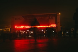 A dimly lit urban scene at night featuring a crowd of people in front of a building. There is a large billboard displaying text in Turkish, and bright red electronic signs illuminate the area, casting a red glow on the pavement.