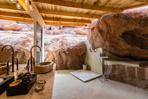 An outdoor bathroom featuring a wooden roof and natural rock walls. It includes a stone sink on a marble countertop with bronze faucets and a modern shower area with a simple platform. Bottles of toiletries are placed on a small side table, and sunlight filters in, enhancing the earthy ambiance.