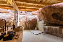 An outdoor bathroom featuring a wooden roof and natural rock walls. It includes a stone sink on a marble countertop with bronze faucets and a modern shower area with a simple platform. Bottles of toiletries are placed on a small side table, and sunlight filters in, enhancing the earthy ambiance.
