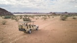 A safari vehicle drives along a dusty path through a desert landscape with scattered bushes and distant hills under a partially cloudy sky. The vehicle is open-roofed and carries several passengers who are seated and observing the surroundings.