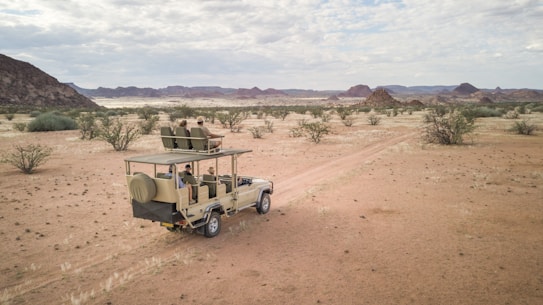 A safari vehicle drives along a dusty path through a desert landscape with scattered bushes and distant hills under a partially cloudy sky. The vehicle is open-roofed and carries several passengers who are seated and observing the surroundings.