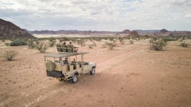 A safari vehicle drives along a dusty path through a desert landscape with scattered bushes and distant hills under a partially cloudy sky. The vehicle is open-roofed and carries several passengers who are seated and observing the surroundings.